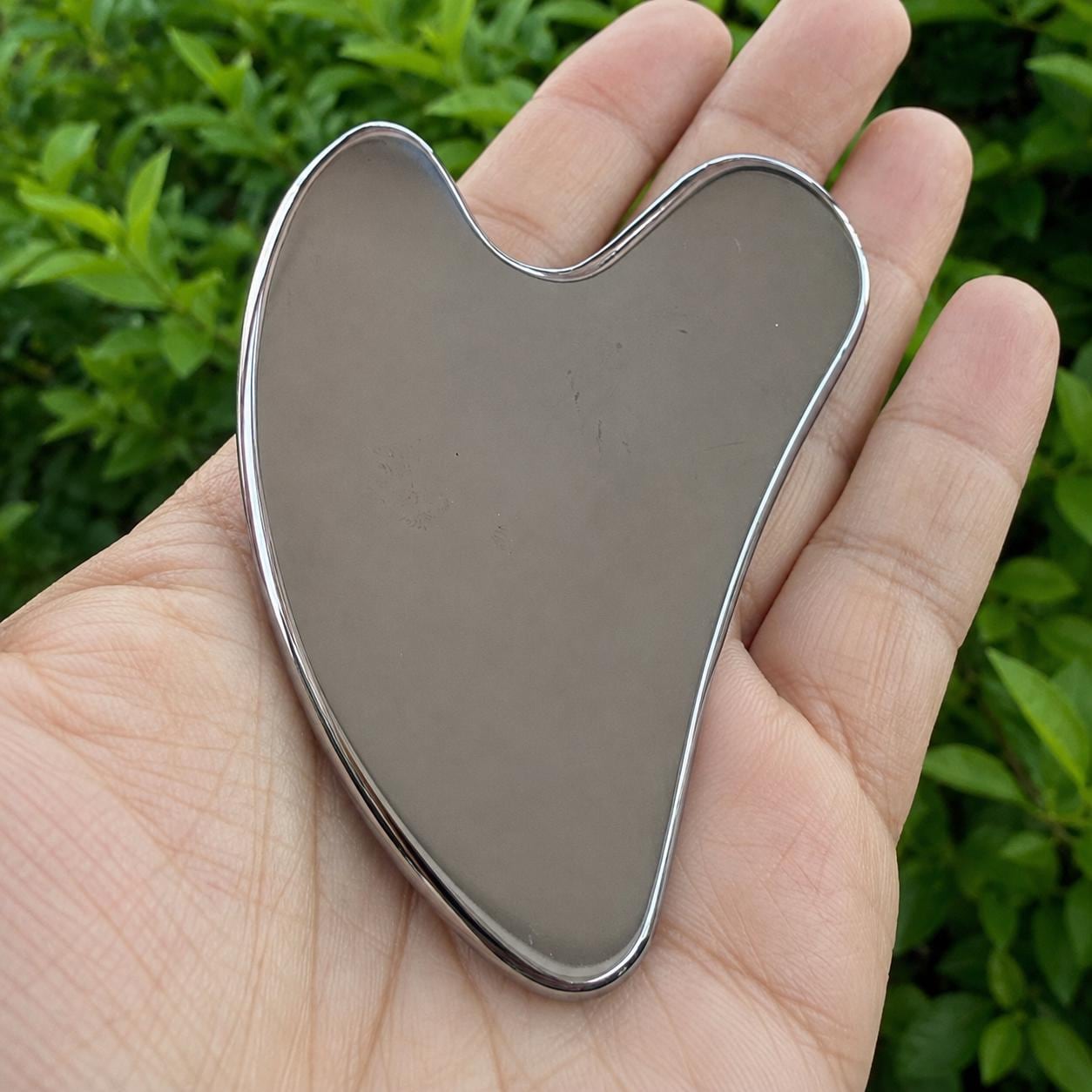 May include: A heart-shaped gua sha tool with a smooth, reflective metallic surface. The object features a curved, ergonomic design with rounded edges and a polished finish, showcasing a solid, uniform grey color throughout its entire structure.
