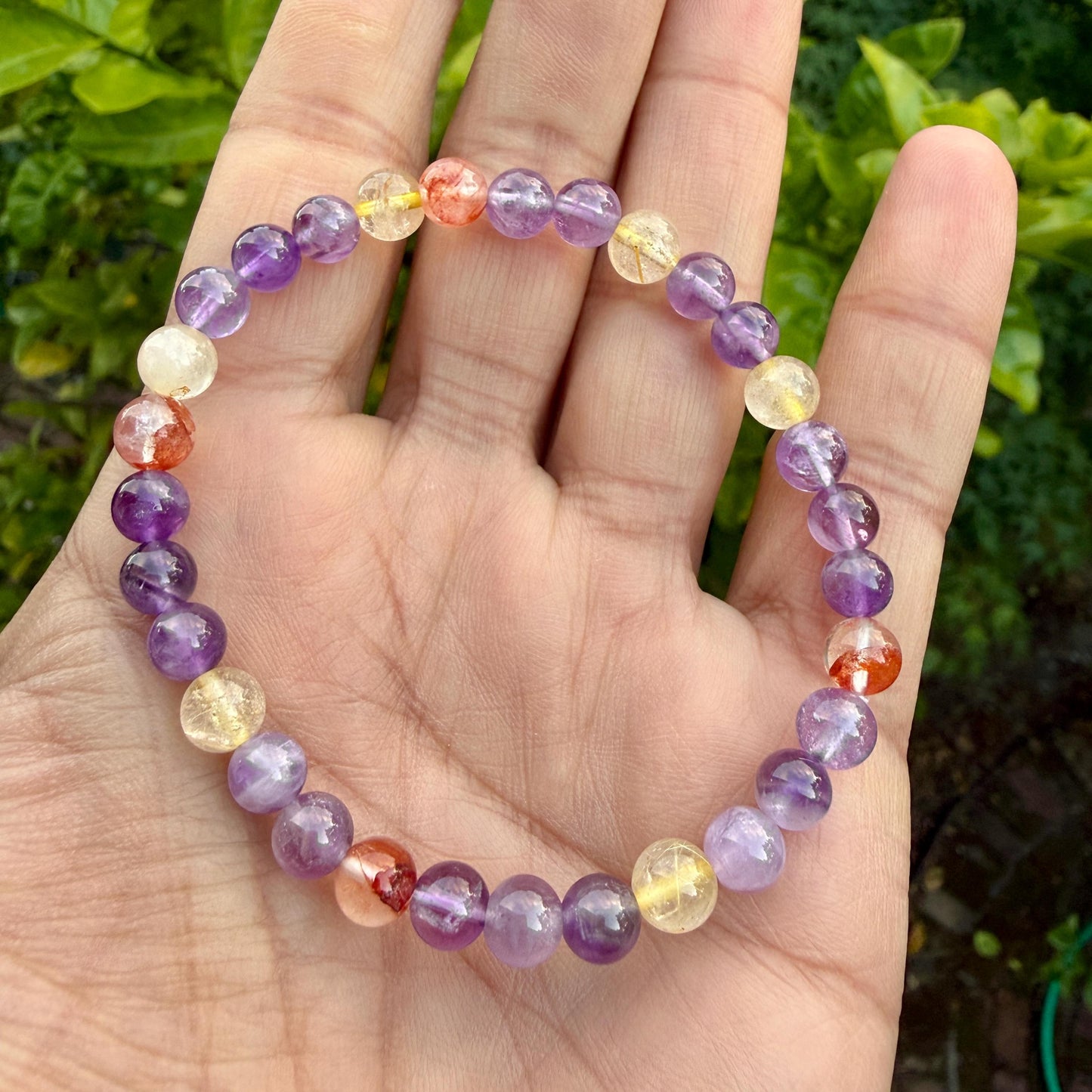 May include: A beaded bracelet featuring a circular arrangement of smooth, polished gemstone beads in shades of purple, translucent yellow, and reddish-orange. The beads are strung together on an elastic cord, creating a continuous, flexible loop.