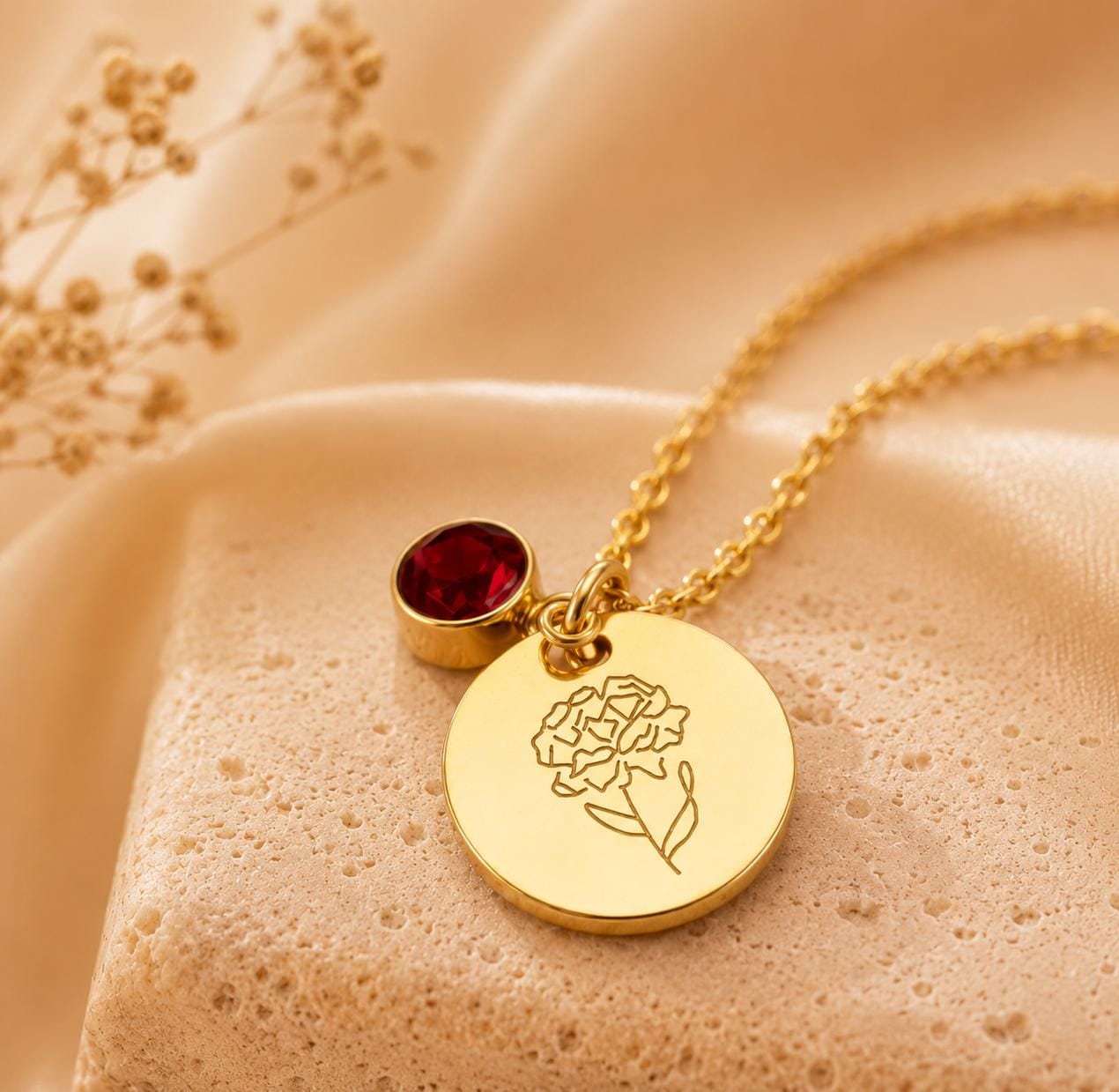 May include: A gold-toned necklace featuring a circular pendant engraved with a line-art flower design and a small, round, deep red gemstone charm. The delicate chain connects to the pendant, which rests on a textured, light-colored surface.