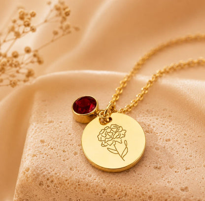 May include: A gold-toned necklace featuring a circular pendant engraved with a line-art flower design and a small, round, deep red gemstone charm. The delicate chain connects to the pendant, which rests on a textured, light-colored surface.