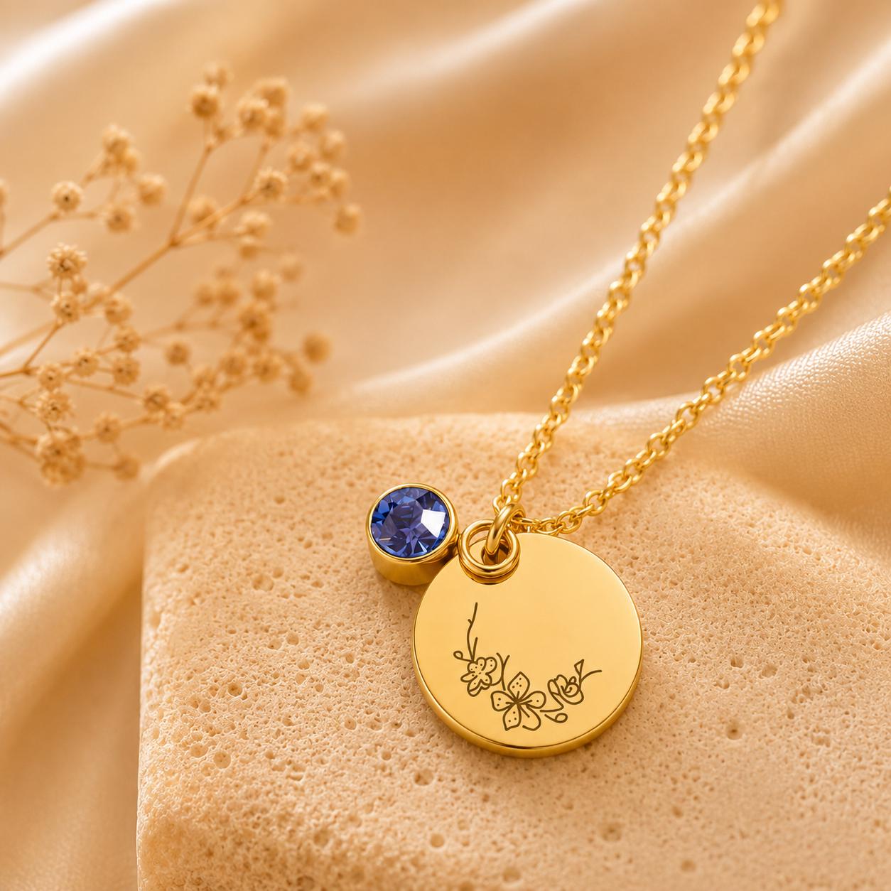 May include: A gold-toned necklace featuring a delicate chain, a circular pendant engraved with a small floral design, and a single round blue crystal charm. The pendant and crystal are suspended together from the chain, creating a layered look.