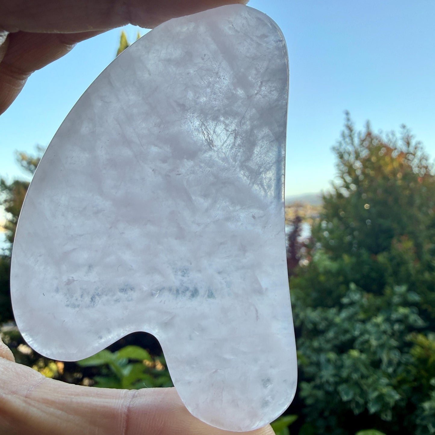 May include: A translucent, pale pink, heart-shaped gua sha tool made of polished quartz stone. The object features smooth, rounded edges and a curved indentation on one side, showcasing a natural, cloudy crystalline texture throughout the surface.