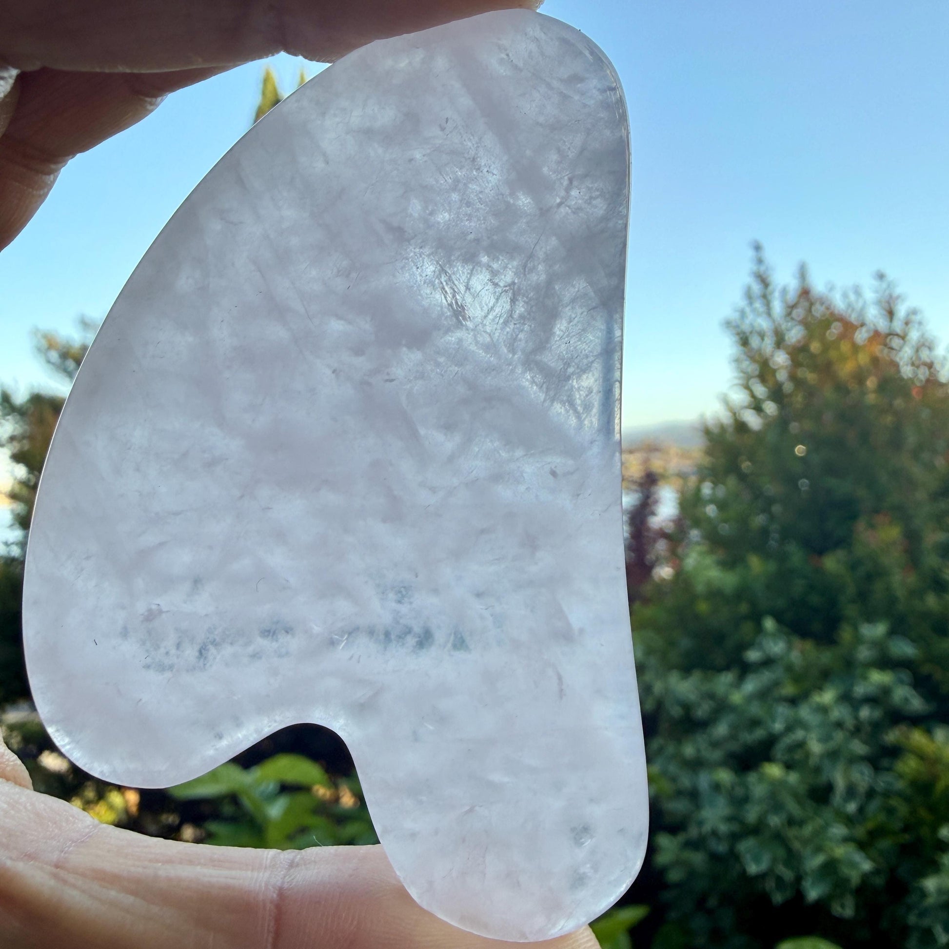May include: A translucent, pale pink, heart-shaped gua sha tool made of polished quartz stone. The object features smooth, rounded edges and a curved indentation on one side, showcasing a natural, cloudy crystalline texture throughout the surface.