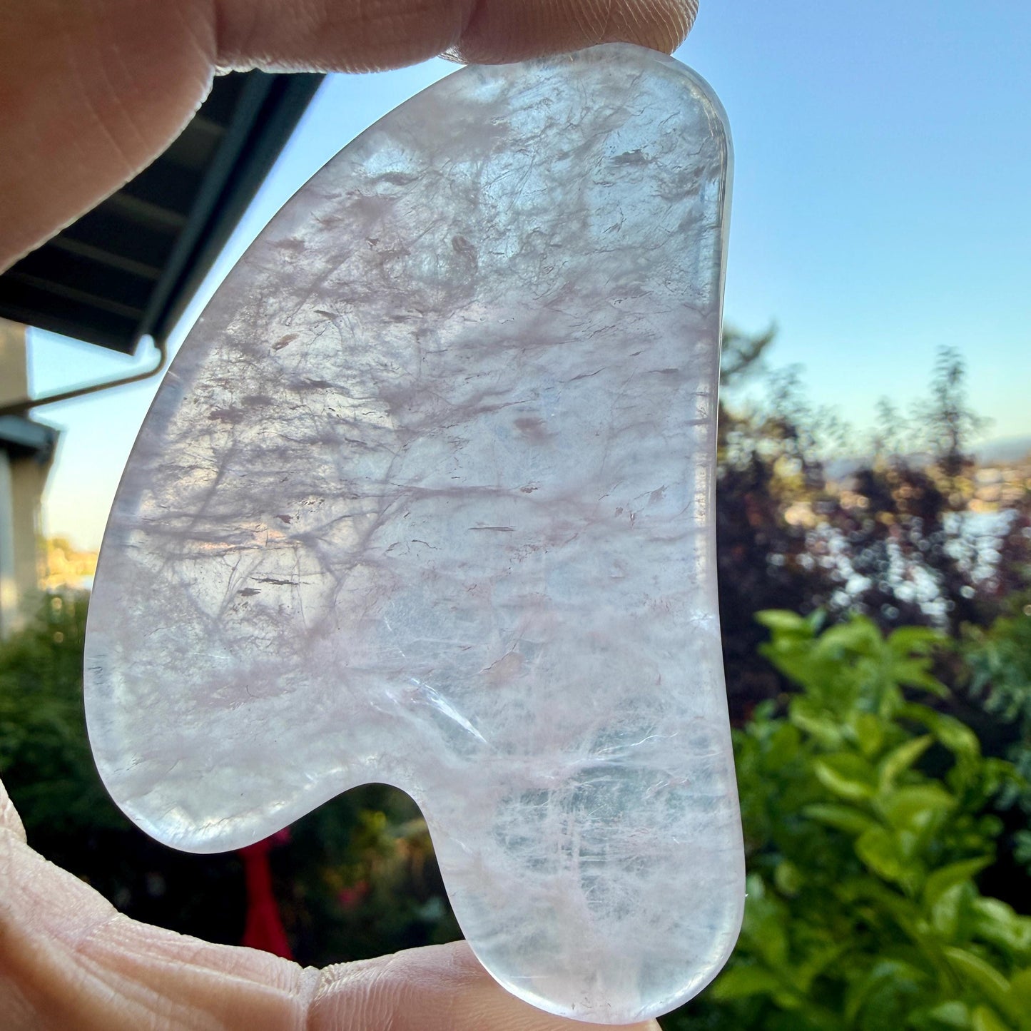 May include: A translucent, pale pink rose quartz gua sha tool with a smooth, polished surface and an irregular, curved shape. The stone features natural internal fractures and cloudy inclusions that catch the light, creating a soft, crystalline appearance.