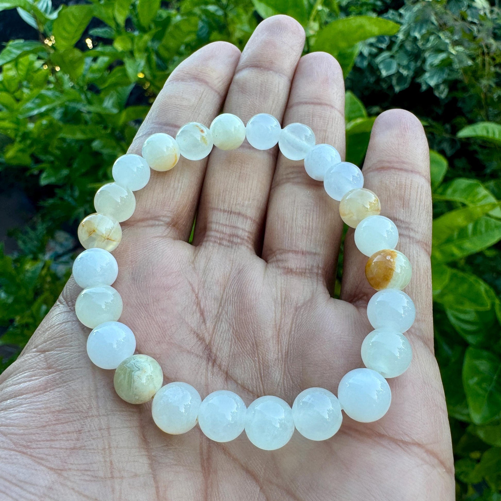 May include: A stretch bracelet composed of smooth, round beads in varying shades of translucent white, pale cream, and soft yellow. The beads are strung together in a circular pattern, displaying a polished finish and subtle internal variations.