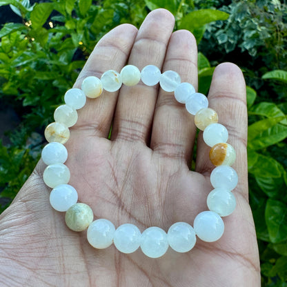 May include: A stretch bracelet composed of smooth, round beads in varying shades of translucent white, pale cream, and soft yellow. The beads are strung together in a circular pattern, displaying a polished finish and subtle internal variations.