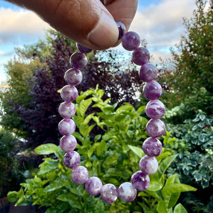 May include: A beaded bracelet composed of round, polished stones in varying shades of purple and white. The beads are strung together in a circular formation, displaying a marbled texture with translucent and opaque sections throughout the piece.