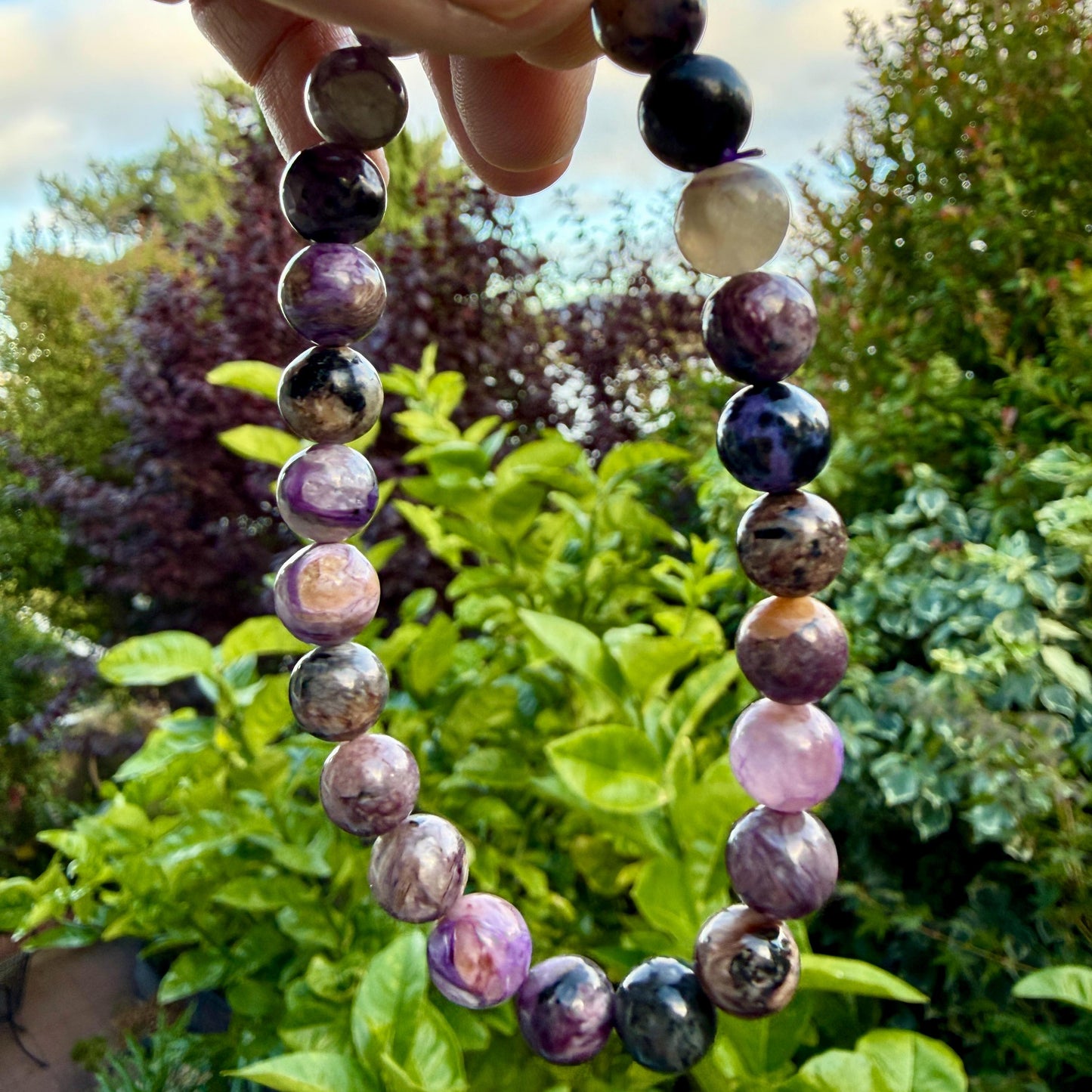 May include: A beaded bracelet featuring round, polished stones in various shades of purple, violet, black, and cream. The beads are strung together in a circular pattern, displaying natural marbling and color variations throughout each individual stone.