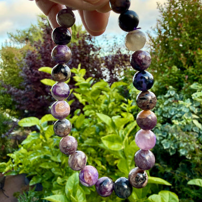 May include: A beaded bracelet featuring round, polished stones in various shades of purple, violet, black, and cream. The beads are strung together in a circular pattern, displaying natural marbling and color variations throughout each individual stone.