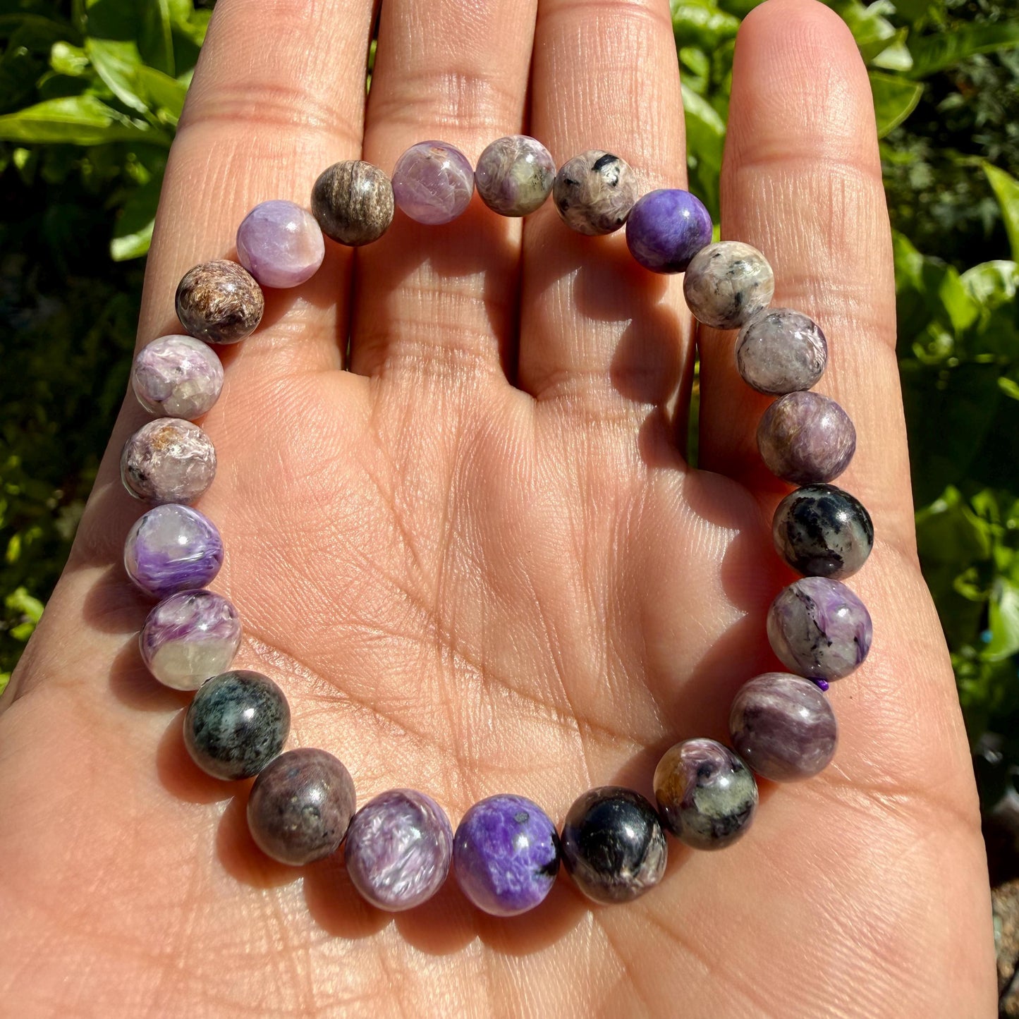 May include: A beaded bracelet featuring round, polished stones in various shades of purple, lavender, gray, and black. The beads are strung together on an elastic cord, displaying natural variations in color, texture, and mineral patterns throughout.