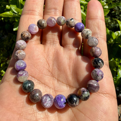 May include: A beaded bracelet featuring round, polished stones in various shades of purple, lavender, gray, and black. The beads are strung together on an elastic cord, displaying natural variations in color, texture, and mineral patterns throughout.
