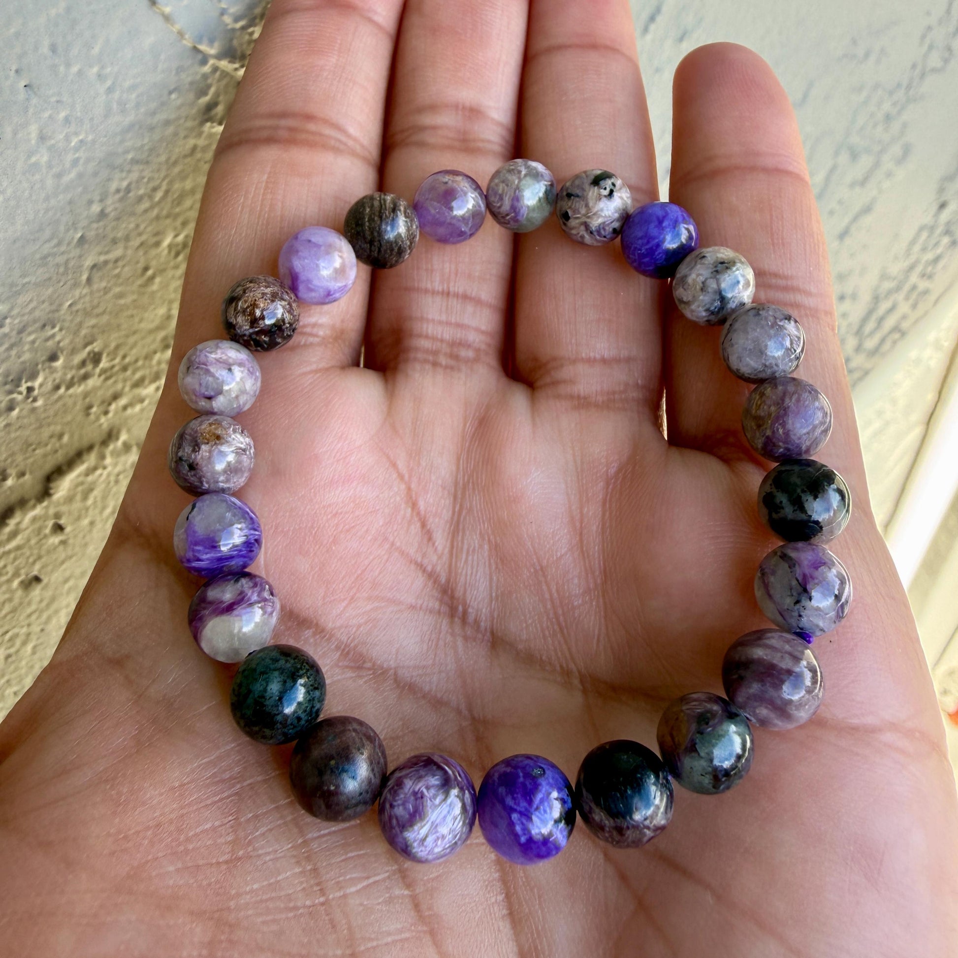 May include: A beaded bracelet featuring round, polished stones in various shades of purple, violet, black, and white. The beads are strung together in a circular pattern, displaying natural marbling and color variations throughout each individual stone.