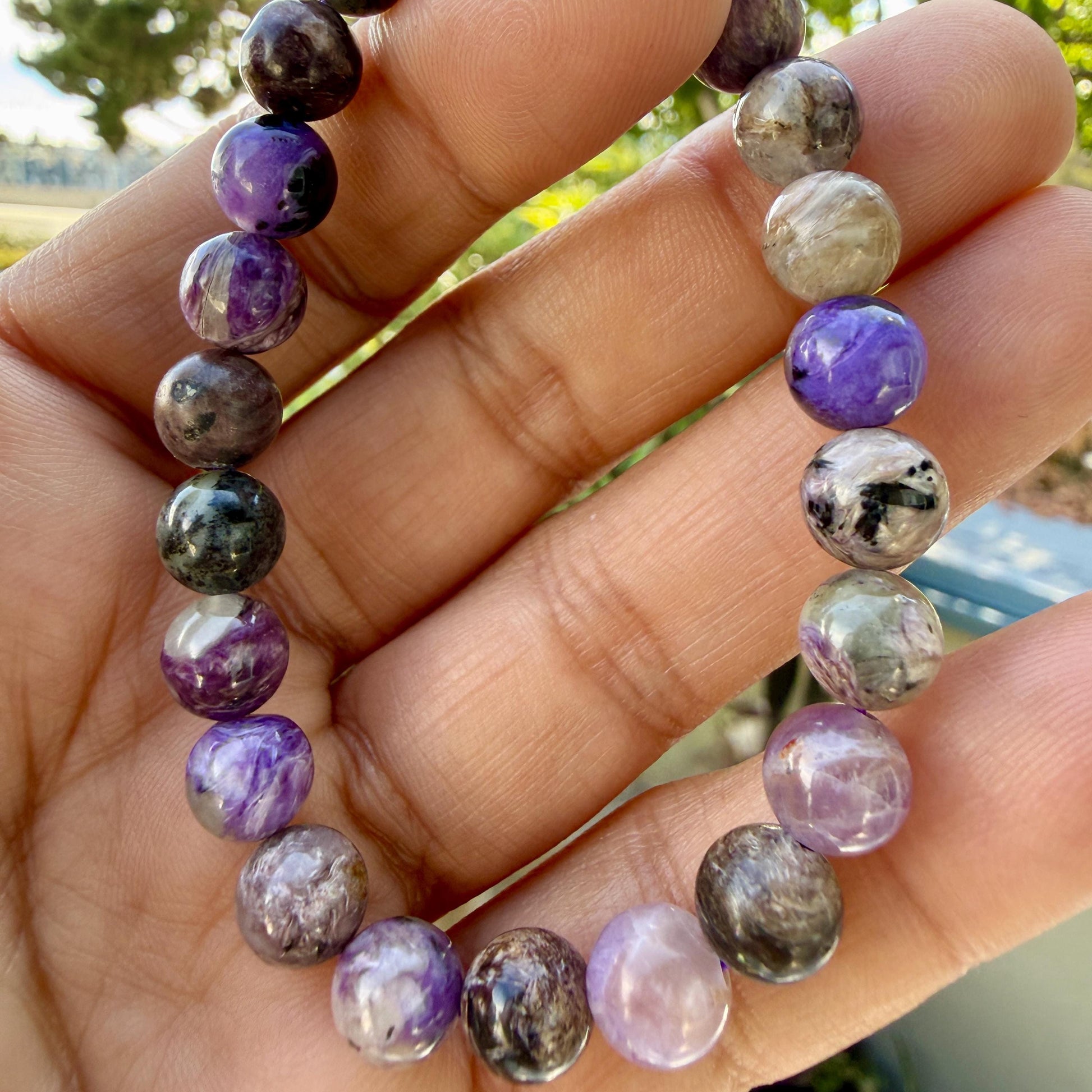 May include: A beaded bracelet featuring smooth, round stones in varying shades of purple, lavender, grey, and black. The beads exhibit natural swirling patterns and glossy finishes, strung together in a circular formation to create a continuous loop.