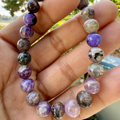 May include: A beaded bracelet featuring smooth, round stones in varying shades of purple, lavender, grey, and black. The beads exhibit natural swirling patterns and glossy finishes, strung together in a circular formation to create a continuous loop.