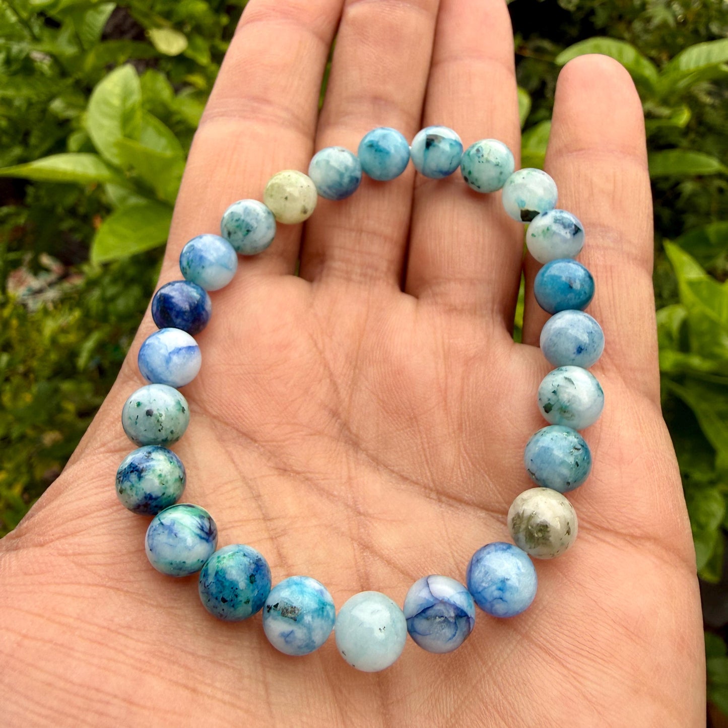 May include: A stretch bracelet composed of round, polished beads in varying shades of blue, white, and light green. The beads feature marbled patterns and translucent qualities, arranged in a circular loop to form a continuous beaded accessory.