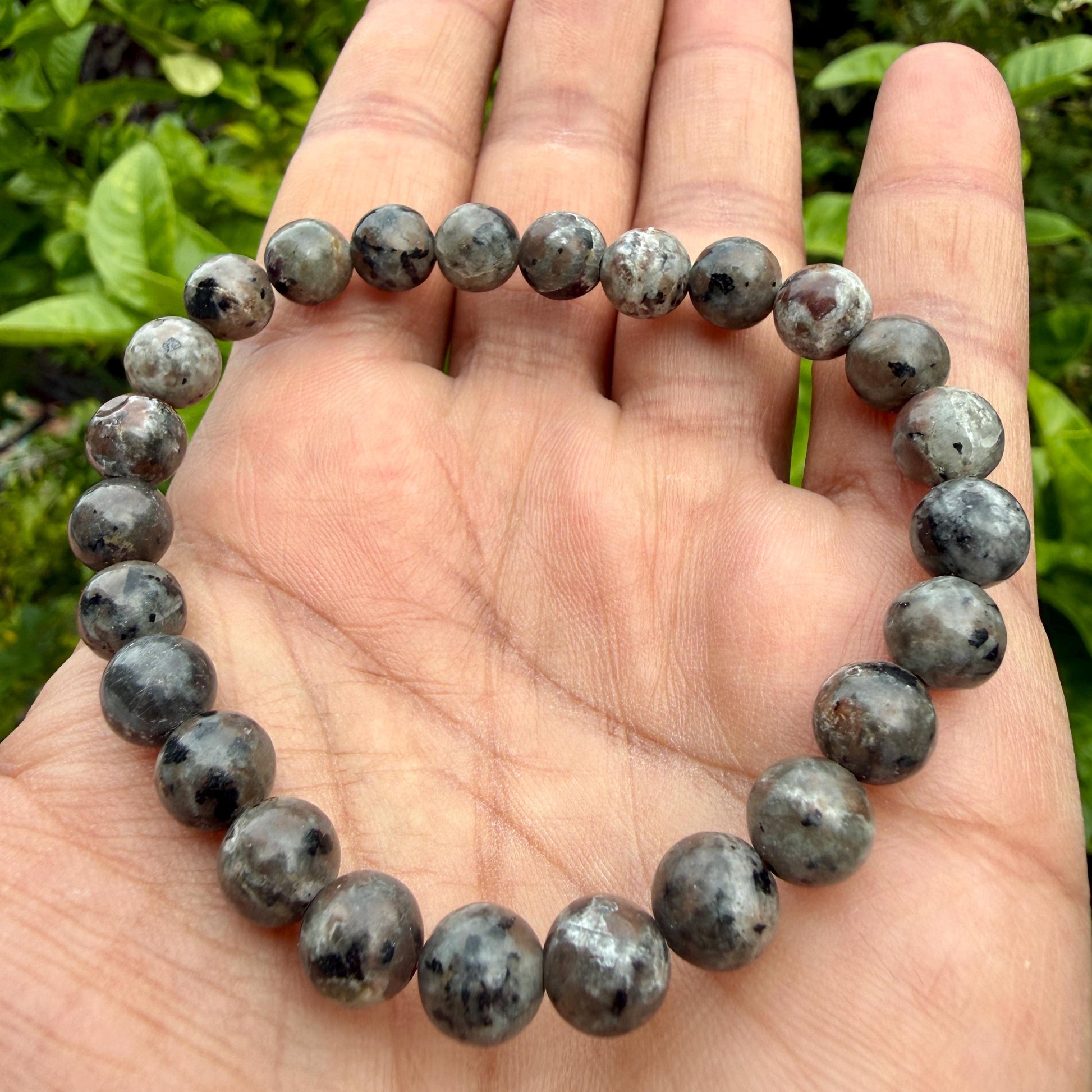May include: A circular beaded bracelet composed of smooth, round, polished stones in shades of grey, charcoal, and muted brown with speckled, mottled patterns throughout each individual bead, strung together on a flexible, elastic cord.