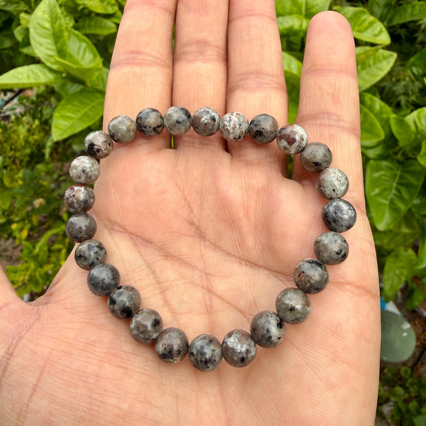 May include: A beaded bracelet composed of round, polished stones in shades of grey, charcoal, and brown. The beads feature a mottled, speckled texture with varying dark and light inclusions throughout the surface, strung together on a flexible cord.
