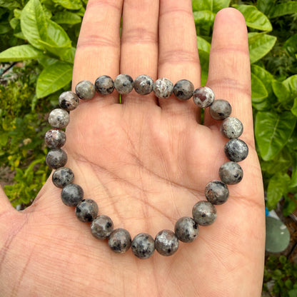 May include: A beaded bracelet composed of round, polished stones in shades of grey, charcoal, and brown. The beads feature a mottled, speckled texture with varying dark and light inclusions throughout the surface, strung together on a flexible cord.