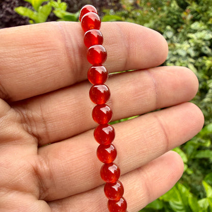 May include: A bracelet composed of a single strand of smooth, round, translucent red carnelian beads. The beads are uniform in size and shape, displaying a vibrant, deep orange-red hue with subtle variations in color intensity across each individual stone.
