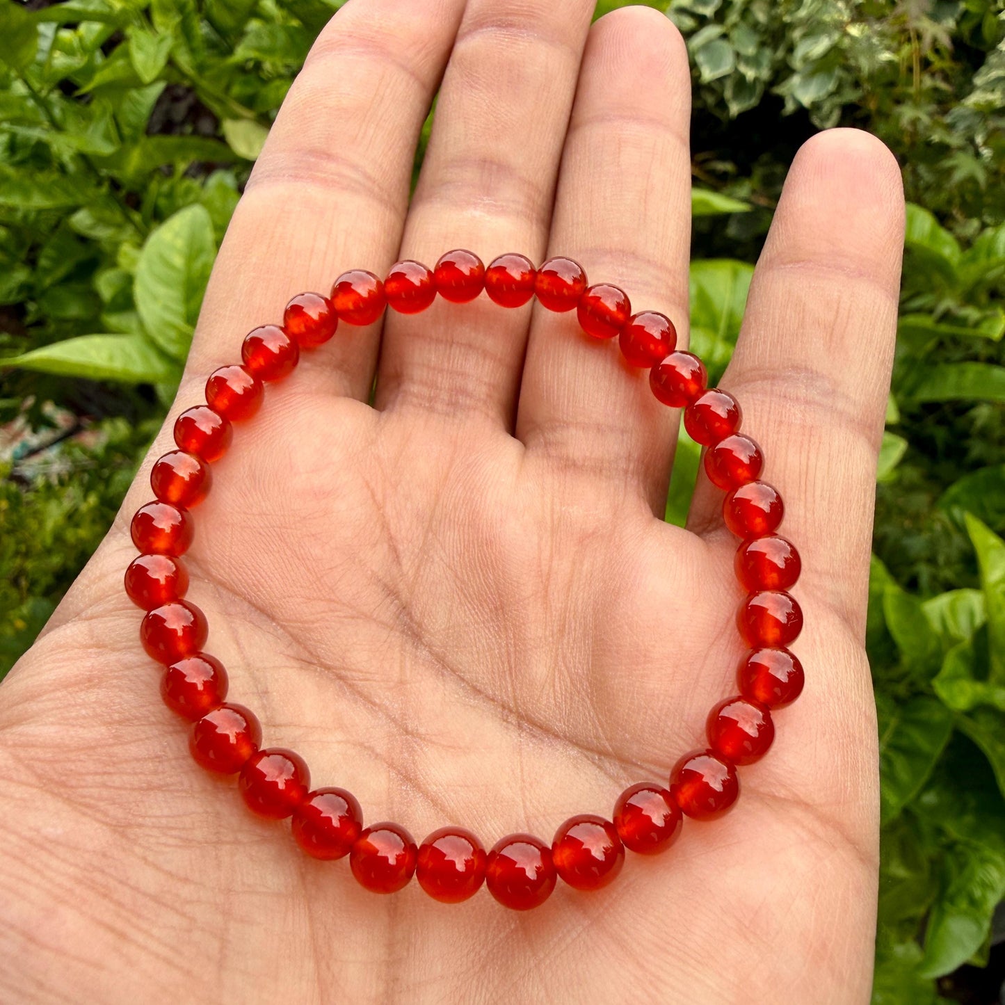 May include: A circular beaded bracelet composed of numerous small, smooth, polished, translucent red gemstone beads strung together on an elastic cord, forming a continuous loop that rests against the palm of a hand.