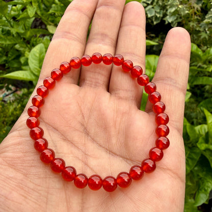 May include: A circular beaded bracelet composed of numerous small, smooth, polished, translucent red gemstone beads strung together on an elastic cord, forming a continuous loop that rests against the palm of a hand.