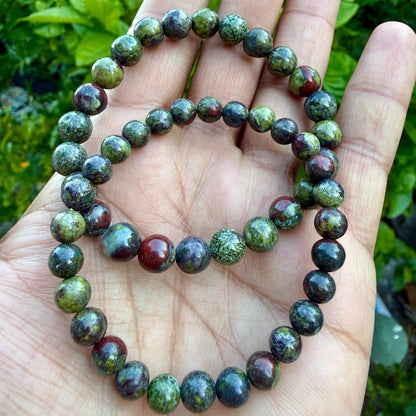 May include: A beaded bracelet featuring round, polished stones in shades of forest green, olive, and deep burgundy. The beads are strung on an elastic cord, creating a circular shape with a smooth, mottled texture across each individual stone surface.