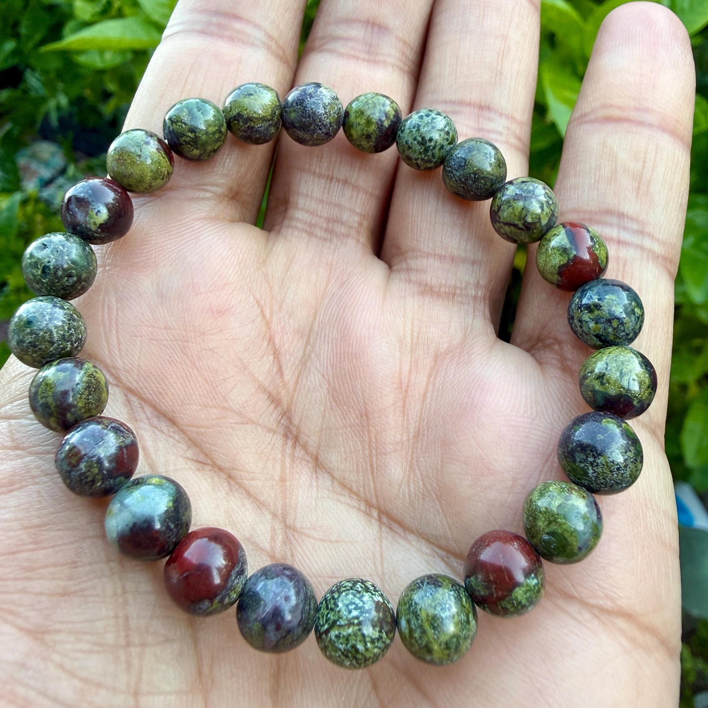 May include: A beaded bracelet featuring round, polished stones in varying shades of olive green, forest green, and deep burgundy red. The beads are strung together in a circular pattern, displaying natural mottled textures and smooth surfaces.