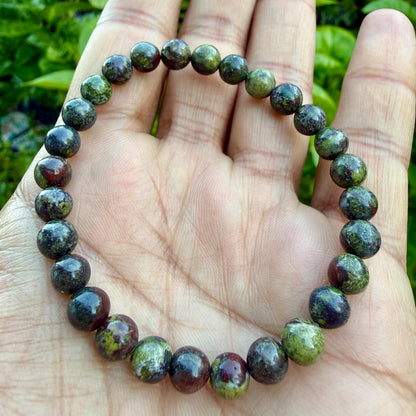 May include: A beaded bracelet composed of round, polished stones in varying shades of deep forest green, olive, and dark burgundy red. The beads are strung together in a circular formation, showcasing natural color variations and smooth textures.