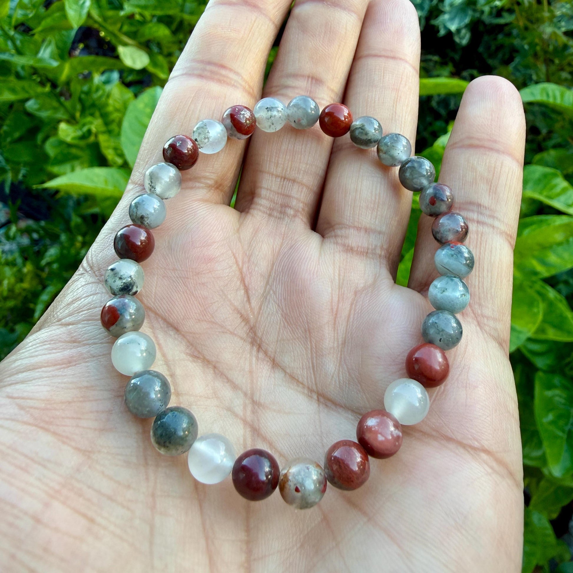 May include: A beaded bracelet featuring alternating round polished stones in shades of deep red, translucent white, and mottled grey. The beads are strung together in a circular pattern, creating a continuous loop of smooth, multi-colored natural stones.