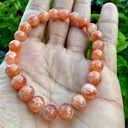 May include: A stretch bracelet composed of round, polished sunstone beads. The beads exhibit a warm, translucent orange hue with subtle, shimmering metallic inclusions throughout the stone, arranged in a continuous circular loop pattern.
