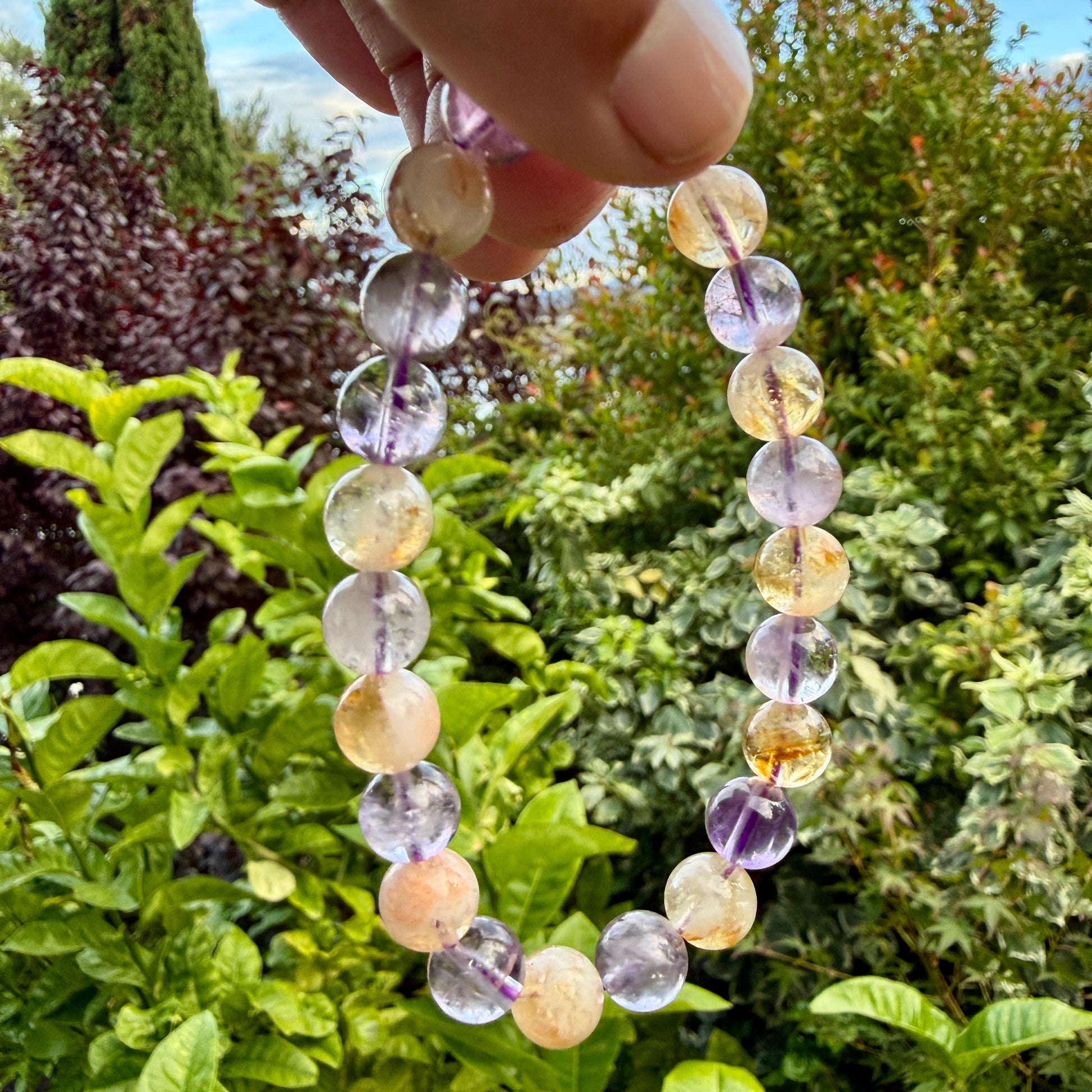May include: A beaded bracelet featuring alternating round, polished gemstones in shades of translucent purple amethyst and warm, golden yellow citrine. The beads are strung on a clear elastic cord, creating a circular shape with varied color tones.
