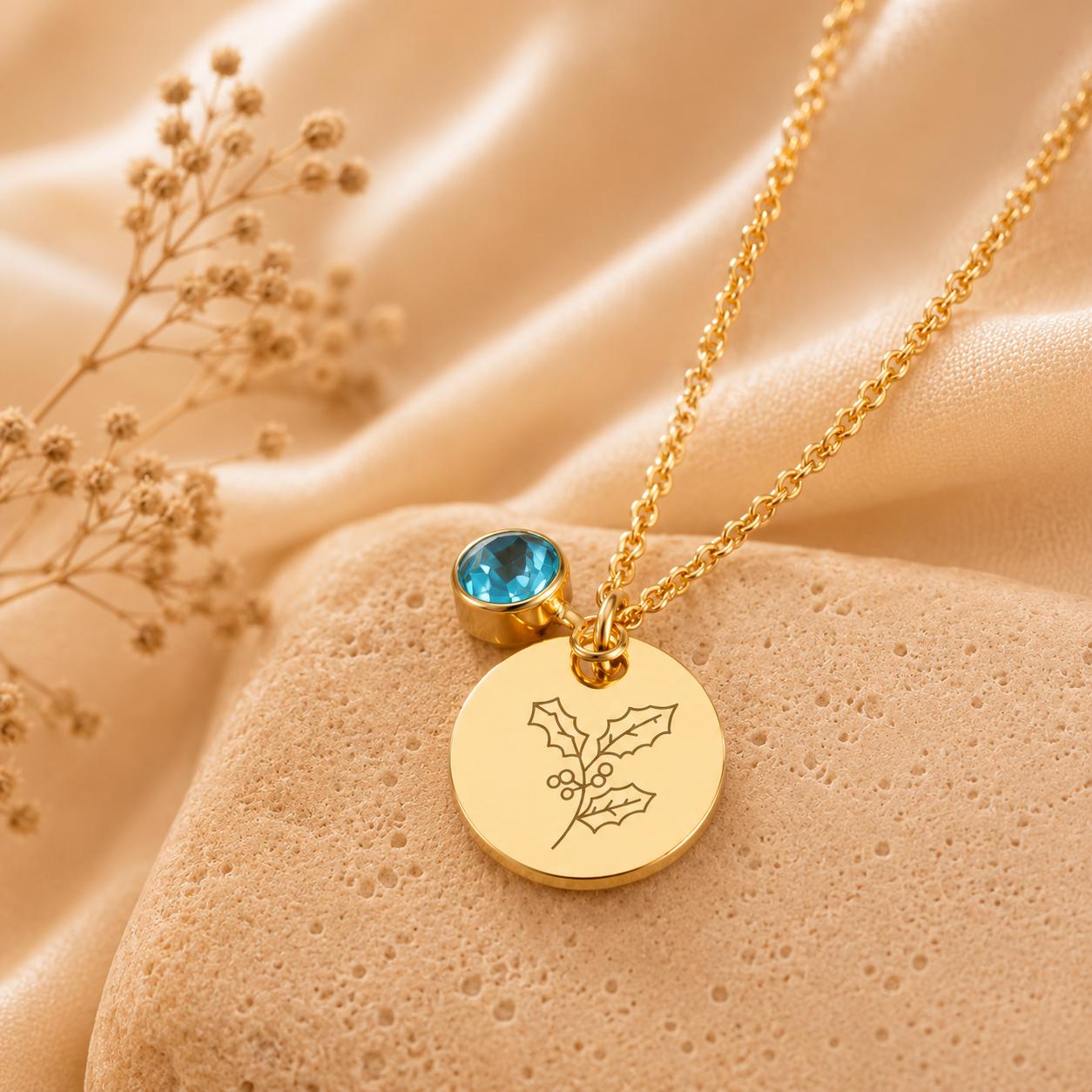 May include: A gold-toned necklace featuring a delicate chain, a round pendant engraved with a line-art holly leaf and berry design, and a small, round, bezel-set blue gemstone charm attached near the top of the pendant.