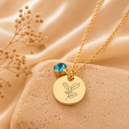 May include: A gold-toned necklace featuring a delicate chain, a round pendant engraved with a line-art holly leaf and berry design, and a small, round, bezel-set blue gemstone charm attached near the top of the pendant.