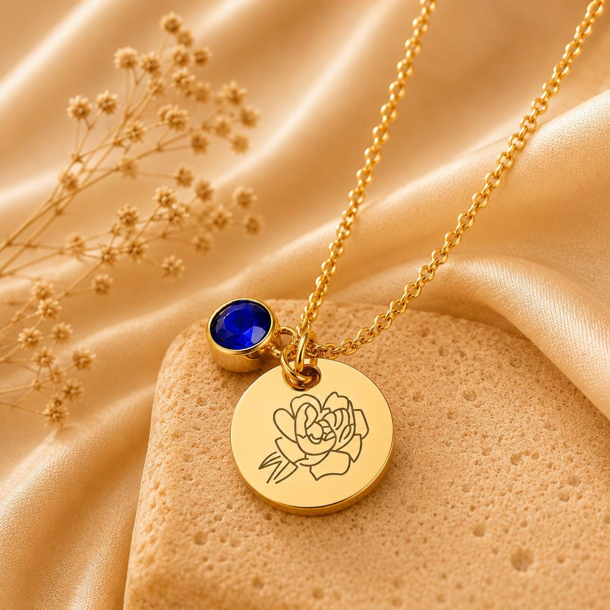May include: A gold-toned necklace featuring a delicate chain, a round pendant engraved with a line-art floral design, and a small, circular, deep blue faceted gemstone charm attached near the top of the pendant.