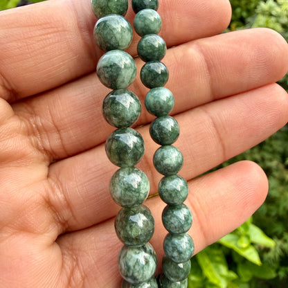 May include: A strand of round, polished green beads with dark mottled patterns and natural veining. The beads vary in size, with one row featuring larger, prominent spheres and the adjacent row consisting of smaller, uniform green stone beads.