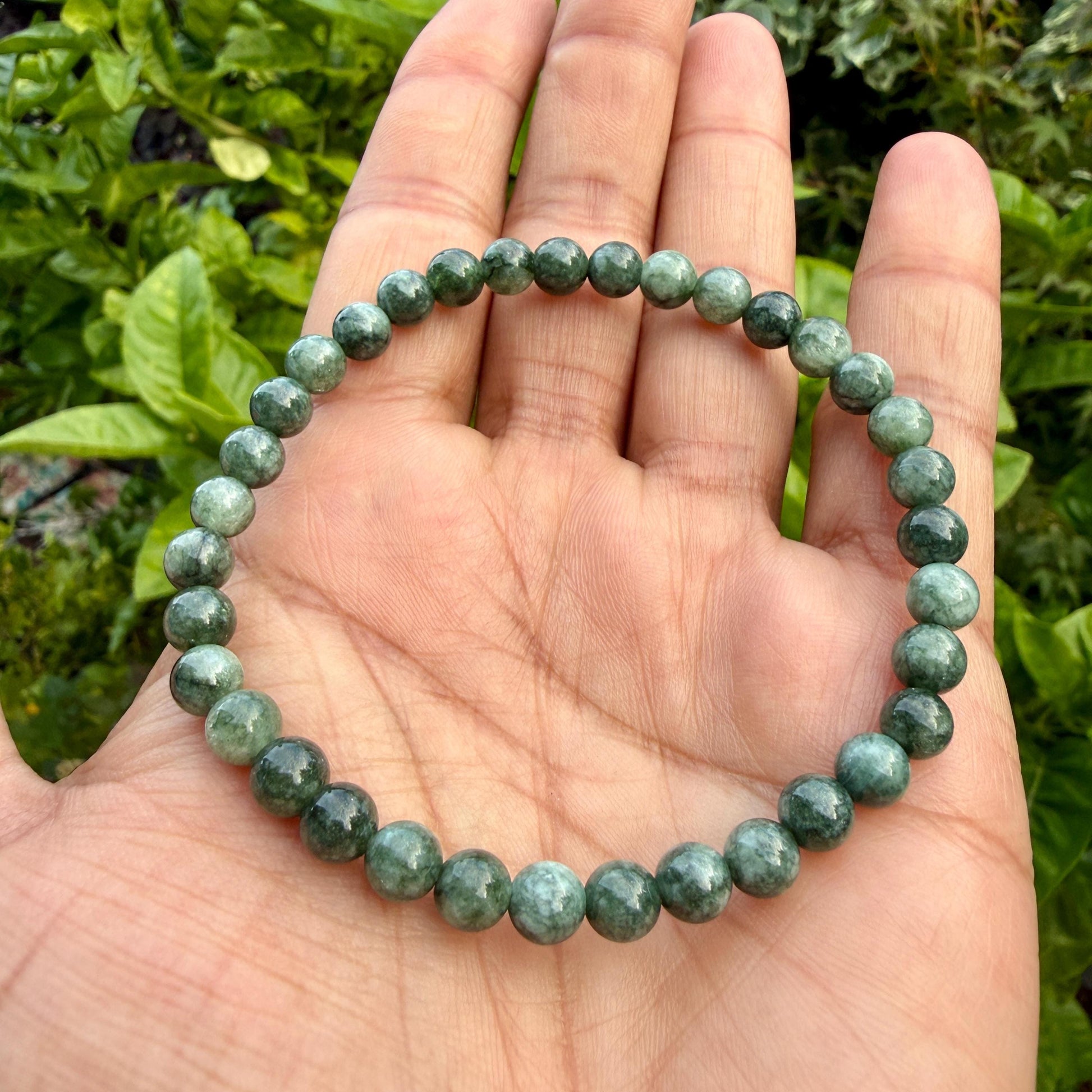 May include: A circular beaded bracelet composed of numerous small, smooth, spherical green stones. The beads exhibit varying shades of deep forest green and lighter sage tones with subtle, natural mottled patterns throughout the surface.