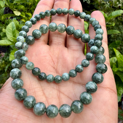 May include: Two circular bracelets composed of polished, spherical beads in varying shades of deep forest green and translucent emerald. The beads feature natural internal marbling and veining, creating a smooth, glossy surface texture throughout.