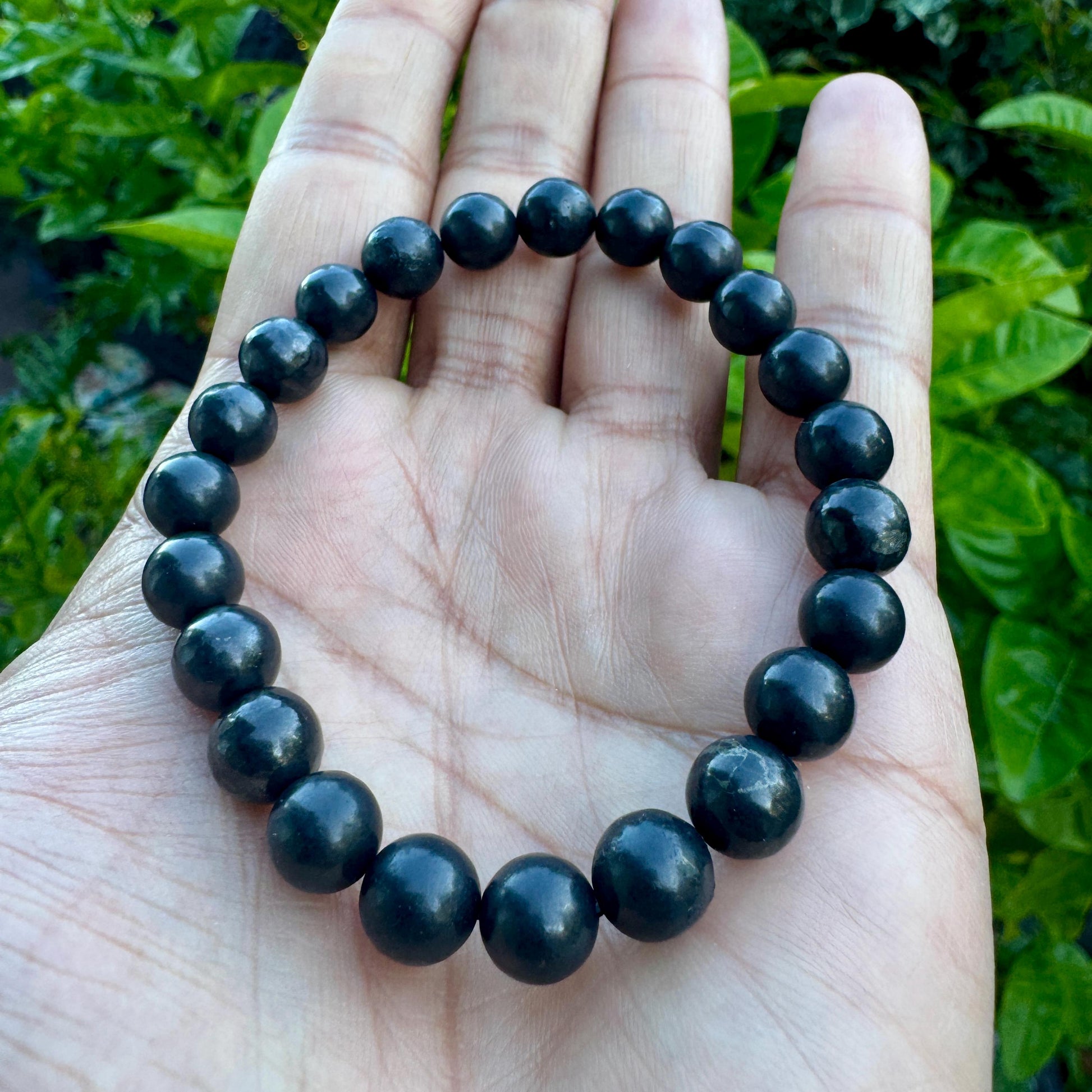 May include: A circular stretch bracelet composed of smooth, round, dark charcoal-gray beads. The beads have a subtle, polished sheen and are uniform in size, strung together to form a continuous loop that rests against the palm of a hand.