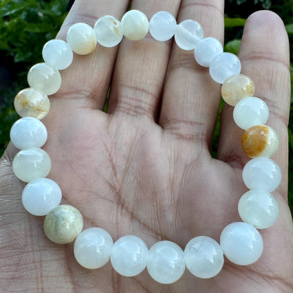May include: A beaded bracelet composed of smooth, round, translucent white stones. Several beads feature subtle, warm golden-yellow inclusions or marbling throughout their surface. The beads are strung together in a circular loop, creating a continuous band.