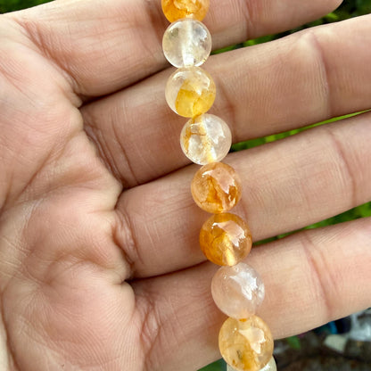 May include: A strand of round, polished beads featuring a mix of translucent clear and vibrant golden-yellow hues. Each bead displays unique internal inclusions and natural variations in color, ranging from pale yellow to deep amber tones.