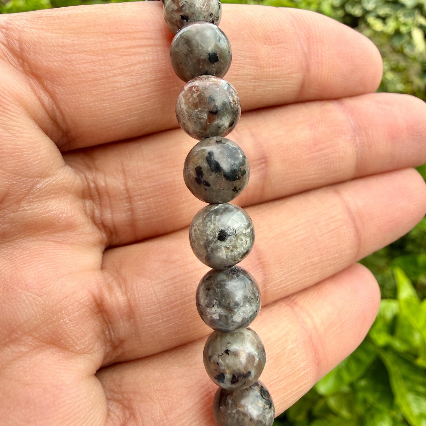May include: A strand of round, polished beads featuring a mottled appearance with shades of grey, brown, and black. Each bead displays unique natural inclusions and dark speckles, creating a varied, earthy texture throughout the entire length.