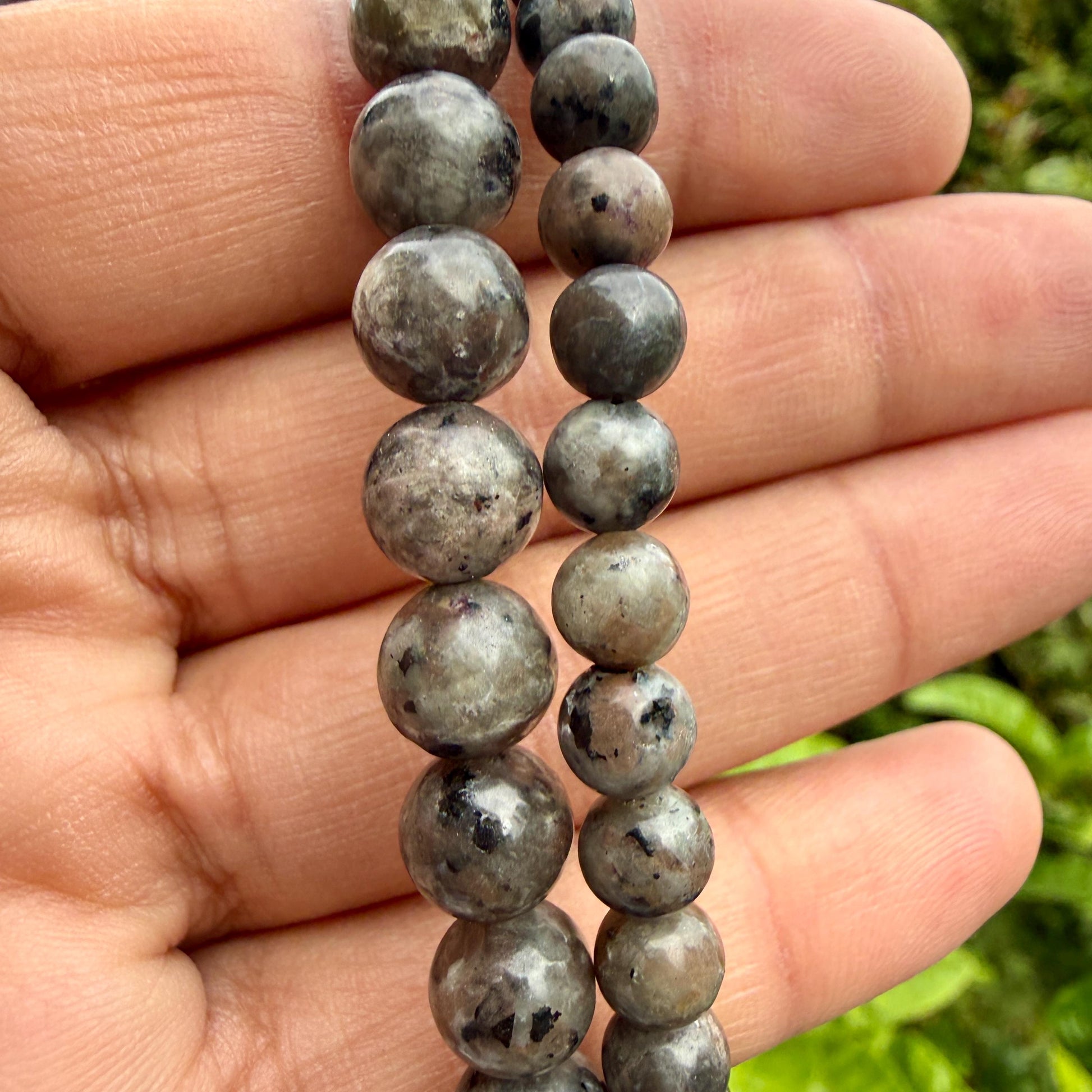 May include: A strand of round, polished beads in shades of gray, brown, and muted green with dark speckled inclusions. The beads vary in size, with one row featuring larger, more prominent spheres and the other row consisting of smaller, uniform beads.