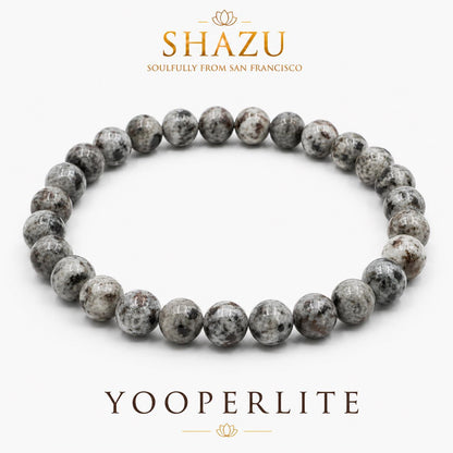 May include: A circular beaded bracelet made of polished Yooperlite stones. The beads feature a mottled pattern of grey, white, and dark brown speckles. The text SHAZU SOULFULLY FROM SAN FRANCISCO and YOOPERLITE is printed in brown above and below the bracelet.