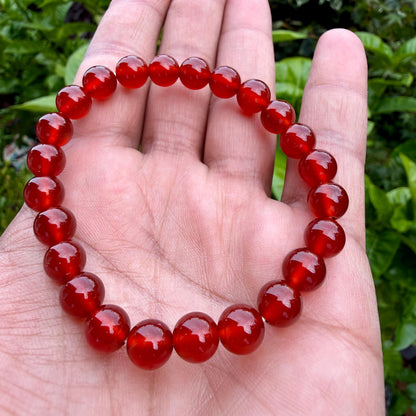 May include: A circular bracelet composed of smooth, polished, translucent red beads. The beads are uniform in size and shape, strung together to form a continuous loop. The deep red color of the stones is consistent throughout each individual bead.