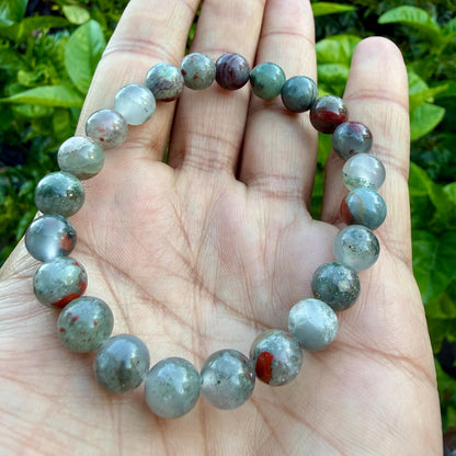 May include: A beaded bracelet featuring round, polished stones in shades of sage green, translucent gray, and deep red. The beads are strung together in a circular pattern, displaying natural variations in color, texture, and internal inclusions.