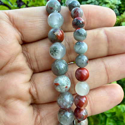 May include: Two strands of round gemstone beads featuring a mix of translucent white, mottled grey, and opaque red stones. The beads vary in size and texture, displaying natural inclusions and color variations throughout the polished spherical surfaces.