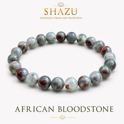 May include: A circular beaded bracelet made of polished African bloodstone spheres. The beads feature a mottled pattern of deep forest green, translucent grey, and opaque dark red inclusions. The text SHAZU SOULFULLY FROM SAN FRANCISCO and AFRICAN BLOODSTONE is visible.