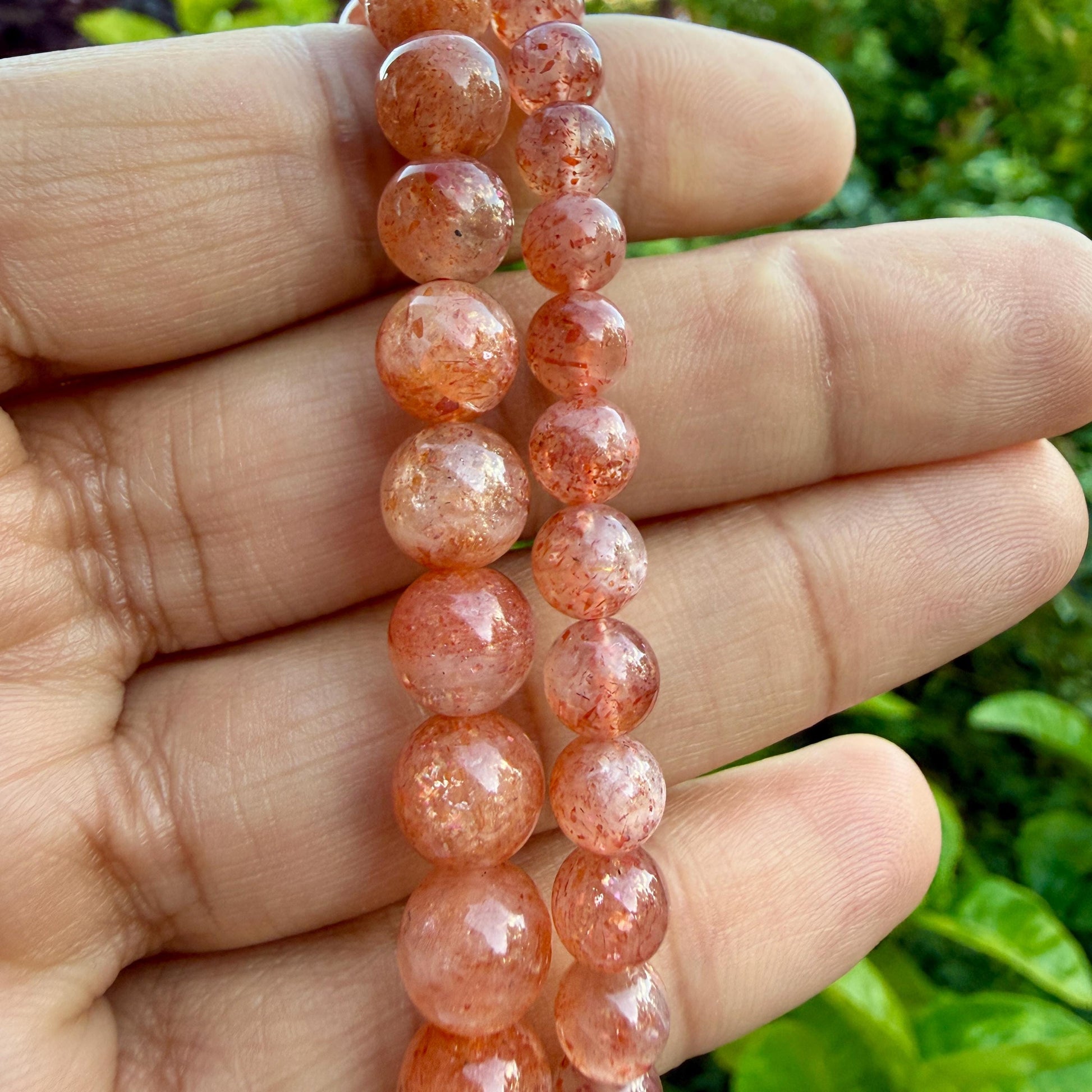 May include: A strand of round, polished sunstone beads featuring a translucent, warm orange-red hue with shimmering, metallic inclusions throughout each stone. The beads are arranged in a vertical line, displaying a natural, speckled, and reflective surface texture.