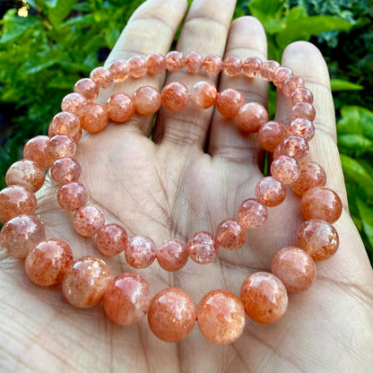 May include: A stretch bracelet composed of round, polished sunstone beads featuring a warm, translucent orange hue with shimmering, glitter-like inclusions throughout each bead, arranged in a single continuous circular strand.