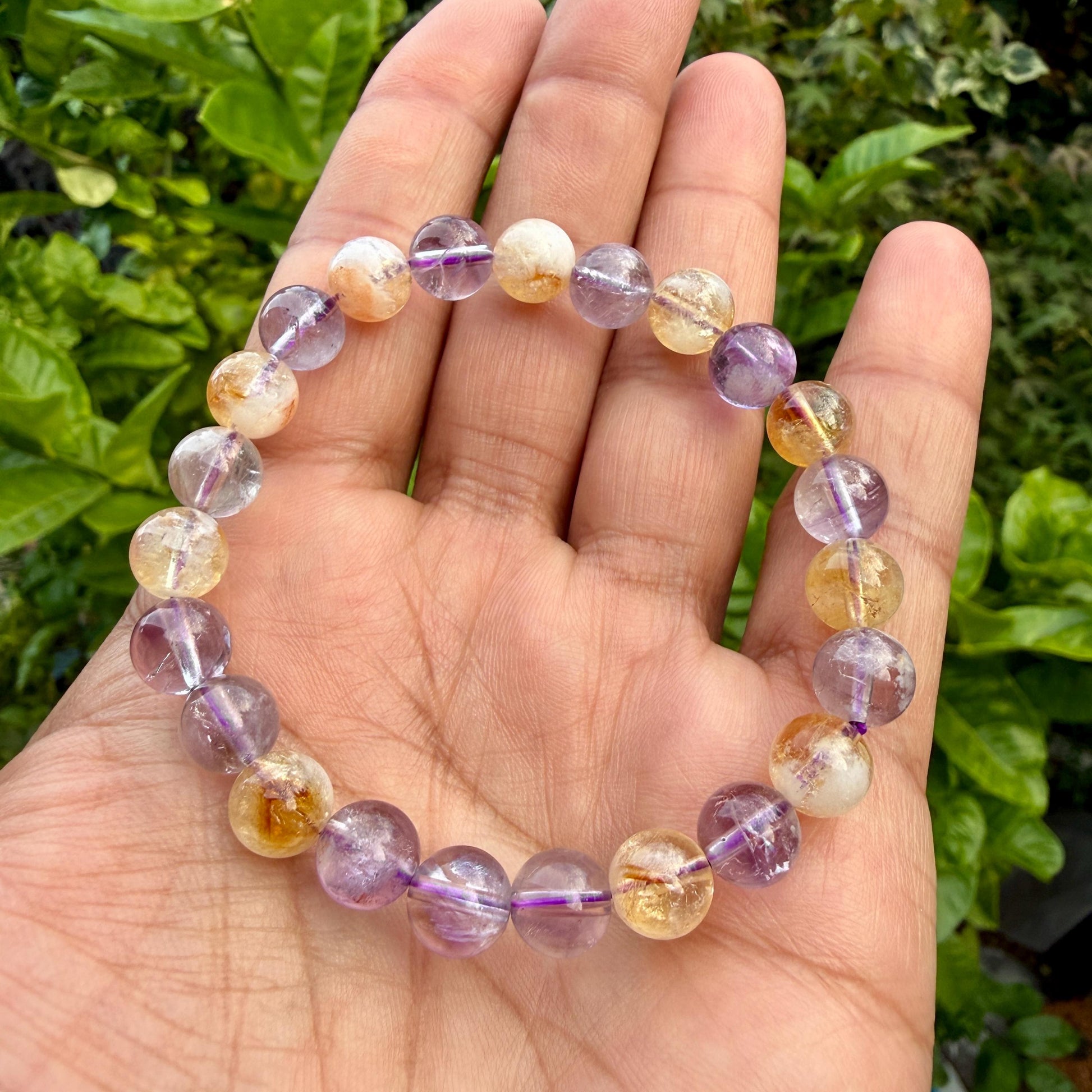 May include: A beaded bracelet featuring alternating round polished gemstones in shades of translucent purple amethyst and golden yellow citrine. The beads are strung on an elastic cord, creating a circular shape with a smooth, reflective surface.